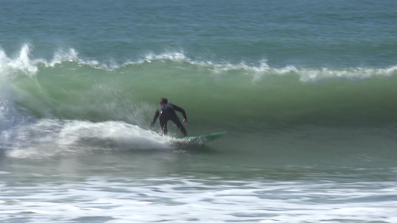 The surfer angles across the same Huntington Beach wave as spray lifts behind the green surfboard and the wave lip starts to thicken overhead.