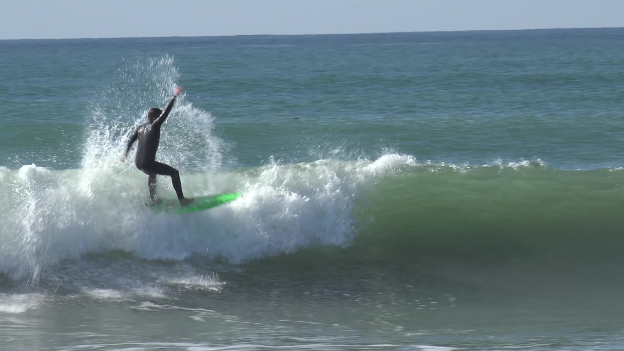A surfer on a green board rises into whitewater at Huntington Beach, California, with spray bursting around the rider and the ocean horizon behind.