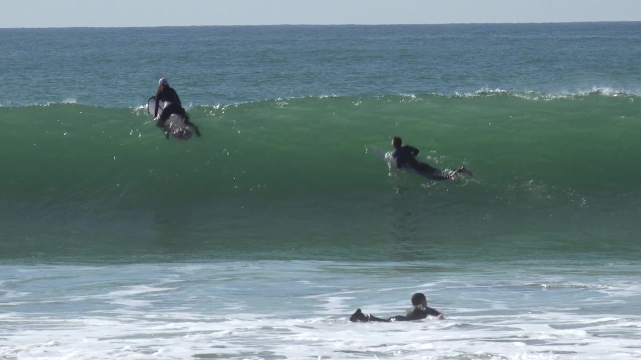 Distant surfers in dark wetsuits face a rising green wave at Huntington Beach, California, with blue ocean and horizon behind them.