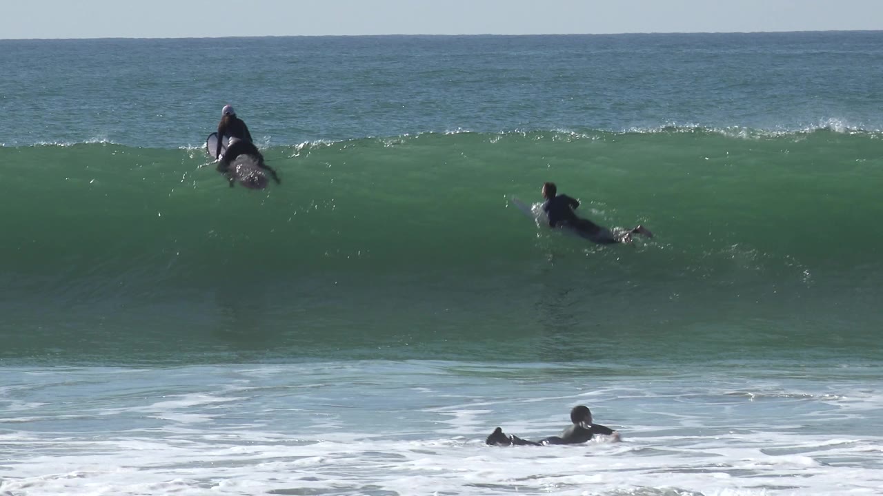 A surfer paddles in the midground while another sits higher on the green face of the Huntington Beach wave under bright daylight.