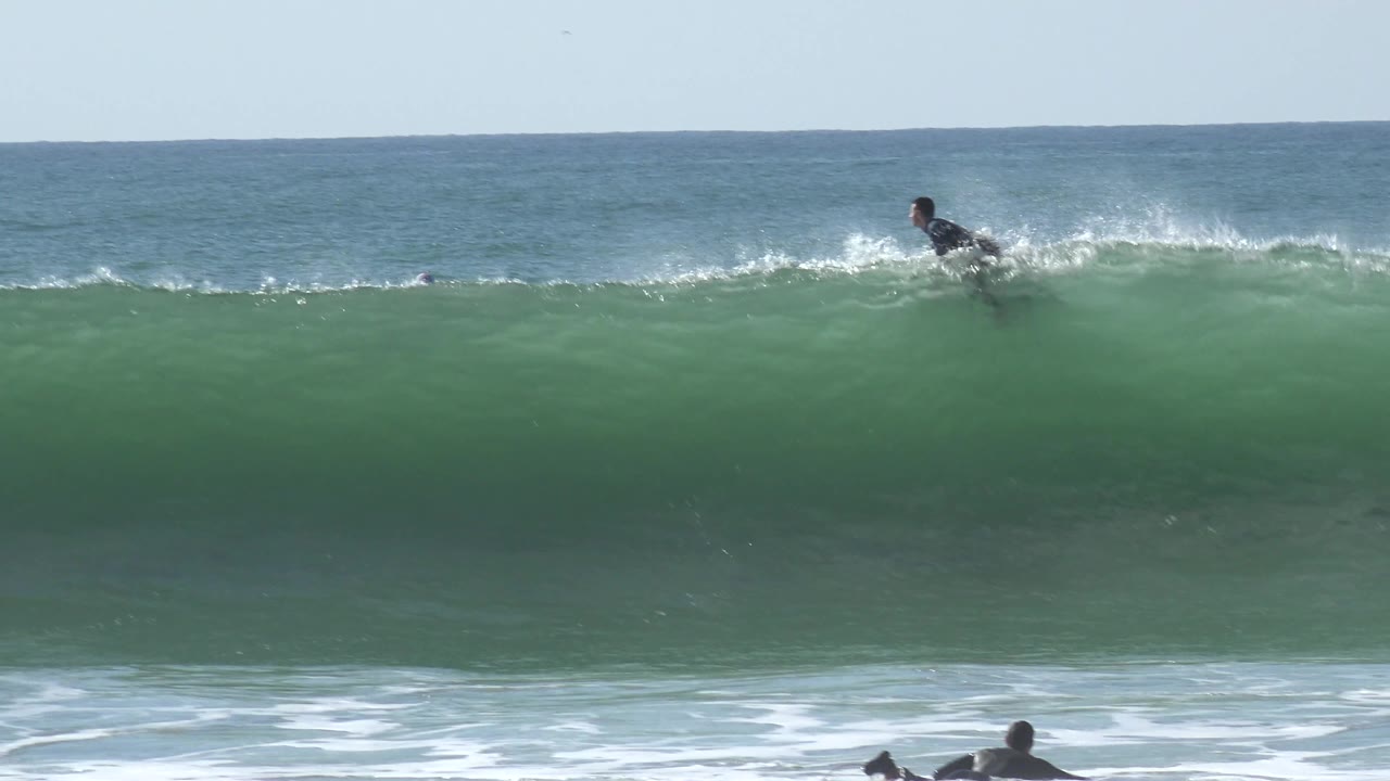 A distant surfer appears near the top of the green wave at Huntington Beach as spray begins to lift from the breaking crest.