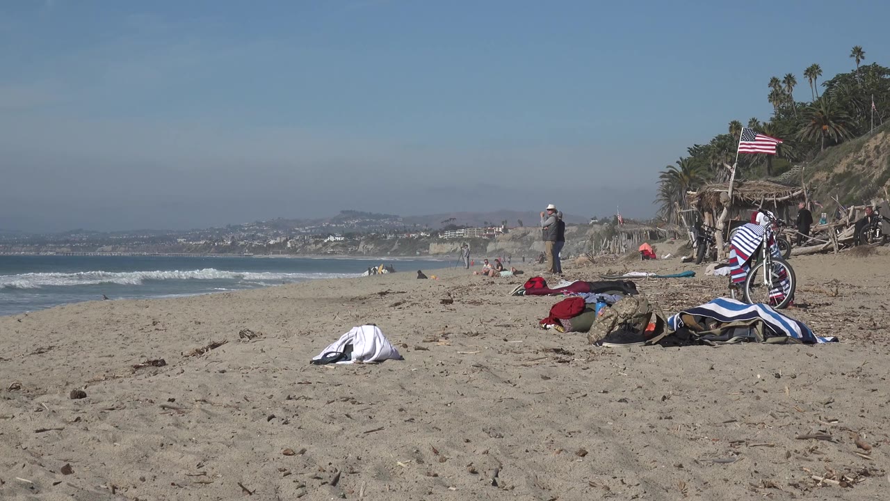 Huntington Beach shoreline view one second in, with the bicycle and towels on the right, a person standing near the waterline, small waves breaking, and the distant coast softened by haze.