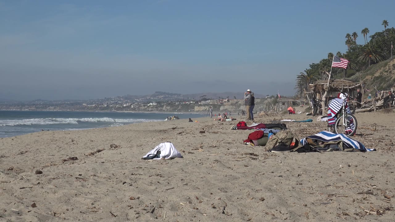 Huntington Beach frame with a low sandy foreground, white towel near center-left, bicycle and striped fabric at right, flag overhead, beachgoers spread near the surf, and a hazy horizon behind them.