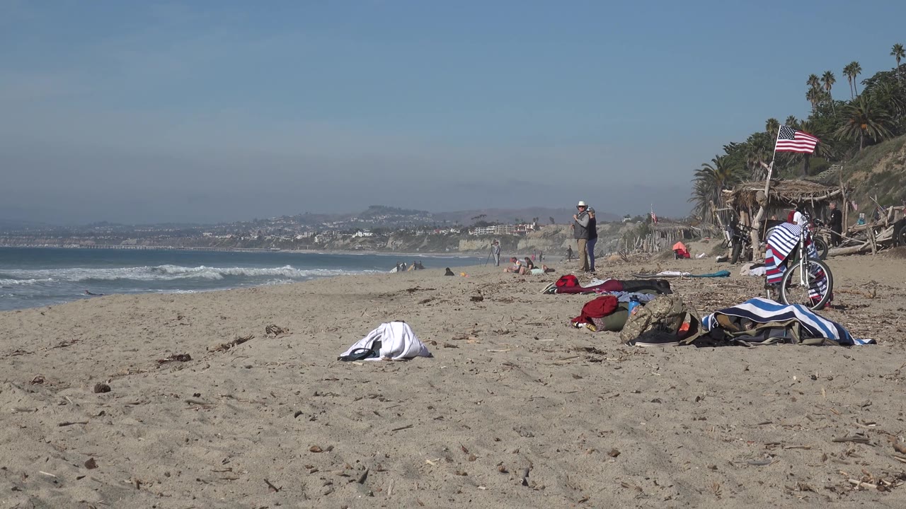 Sandy beach foreground with bicycle, towels, flag, ocean, and distant people.