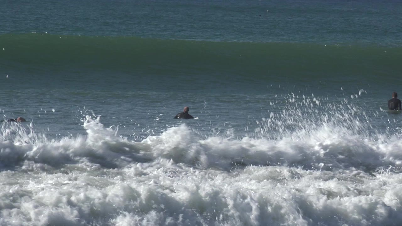 At Huntington Beach, California, several distant wetsuited surfers sit or paddle beyond foamy shorebreak while a dark green wave builds across the background.