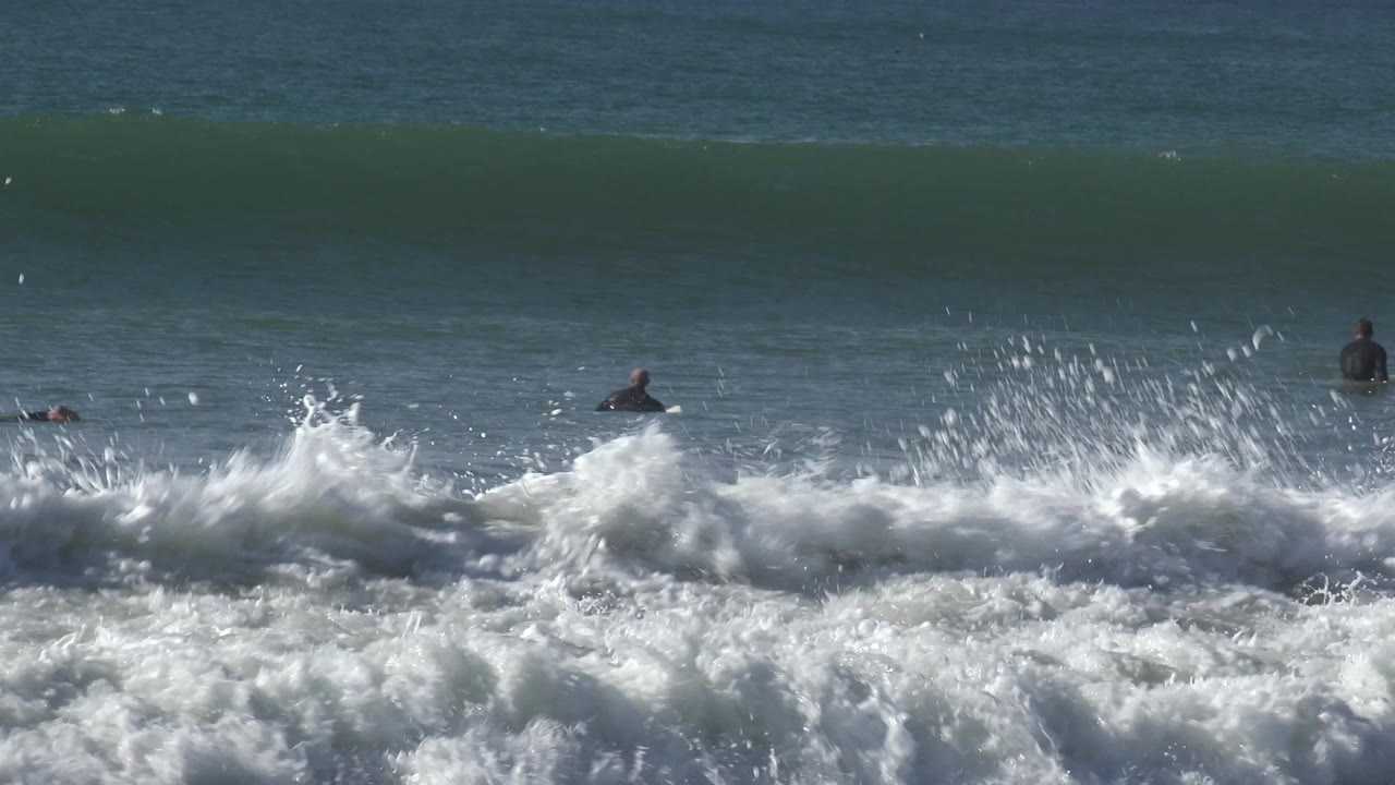 White surf explodes in the foreground with surfers farther out in the water.