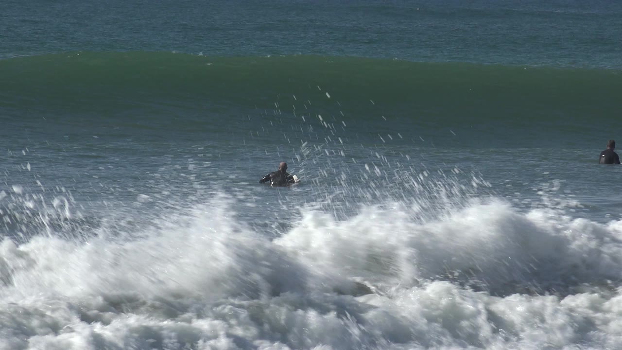 At Huntington Beach, California, a broad green wave forms a smooth horizontal wall behind distant surfers floating in calmer midground water.