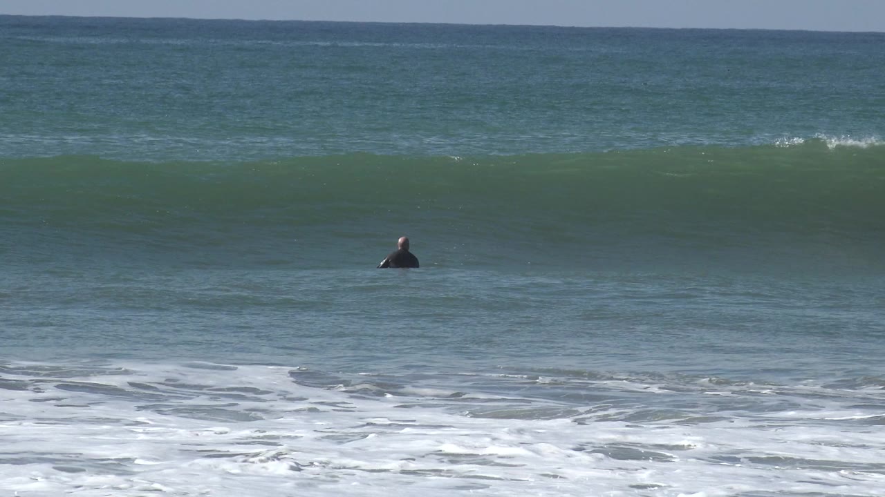 At Huntington Beach, a small distant surfer sits in blue-green water while a broad wave rises behind white shore foam.
