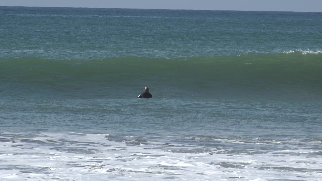 At Huntington Beach, a distant surfer appears near the center of frame below a smooth green wave with textured blue water beyond.