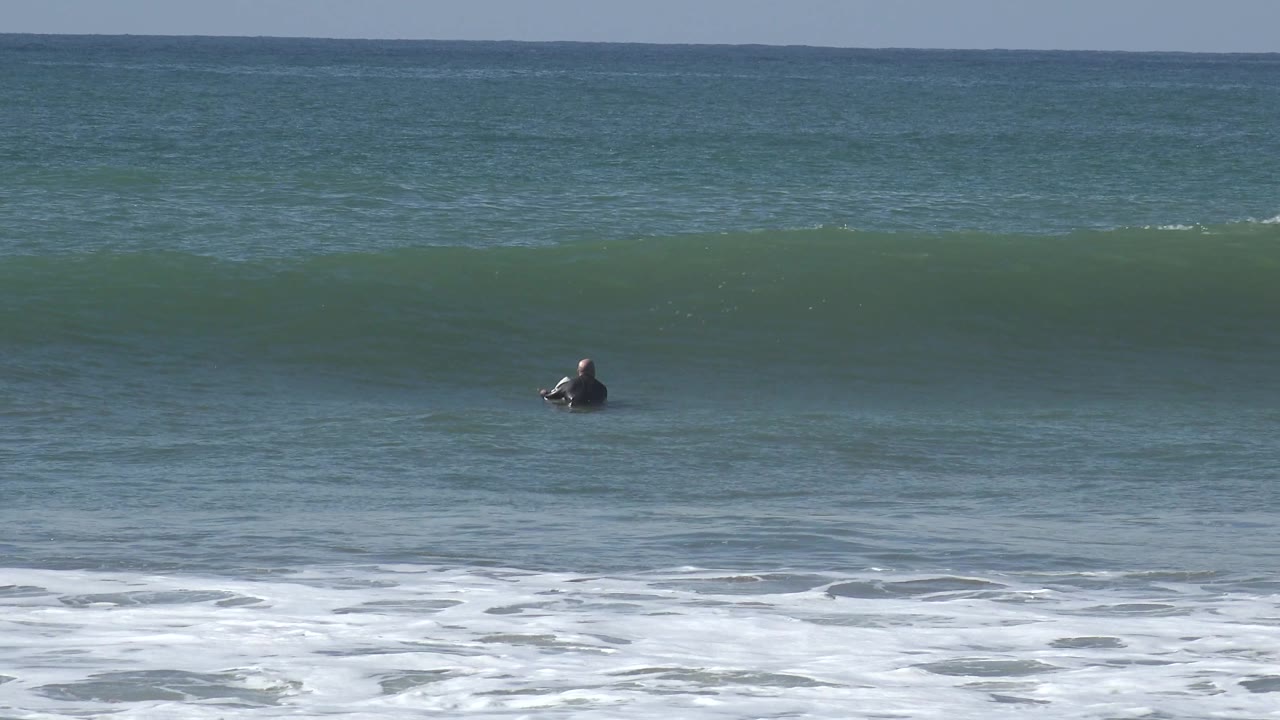 At Huntington Beach, a small distant surfer sits toward the right side as a low green wave stretches across the frame.