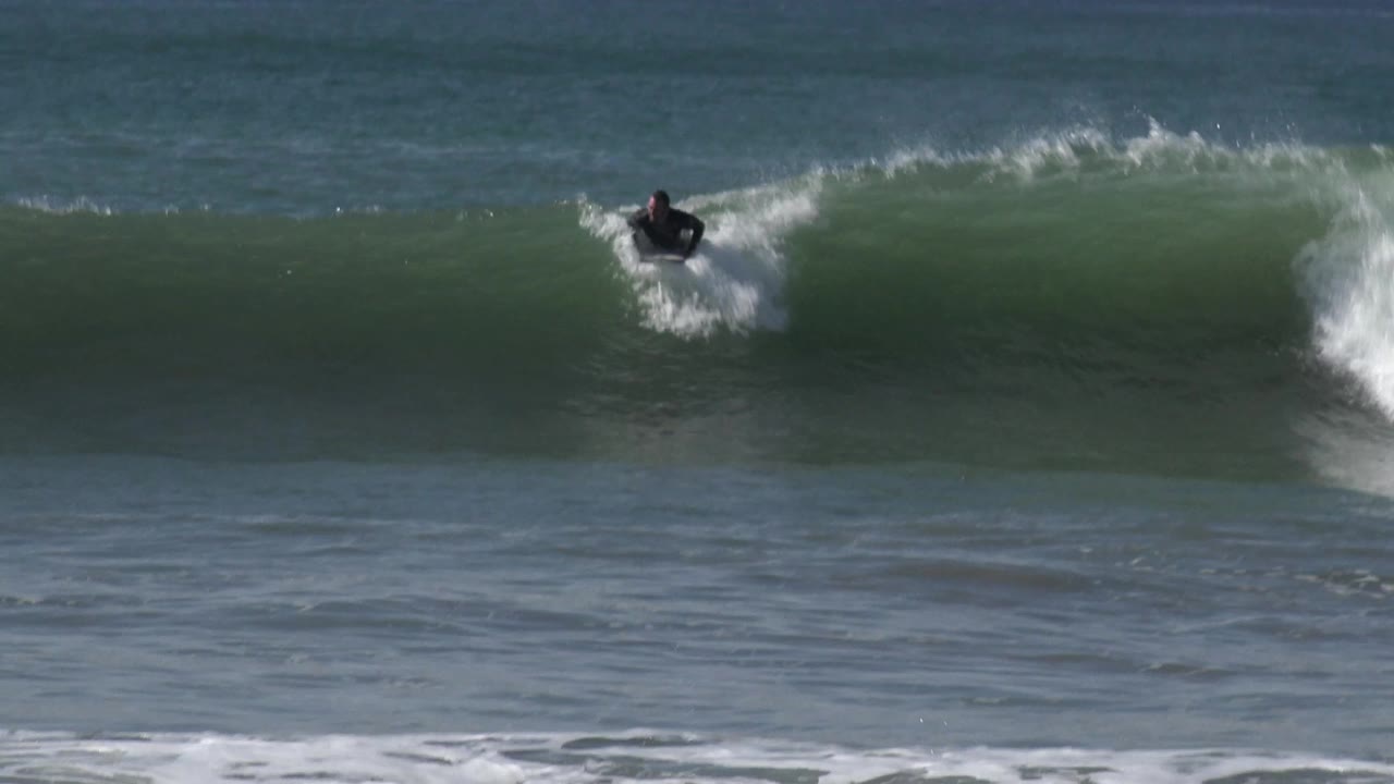 A surfer in a black wetsuit rides near the top of a green wave at Huntington Beach, with white spray breaking behind him and open ocean on the horizon.