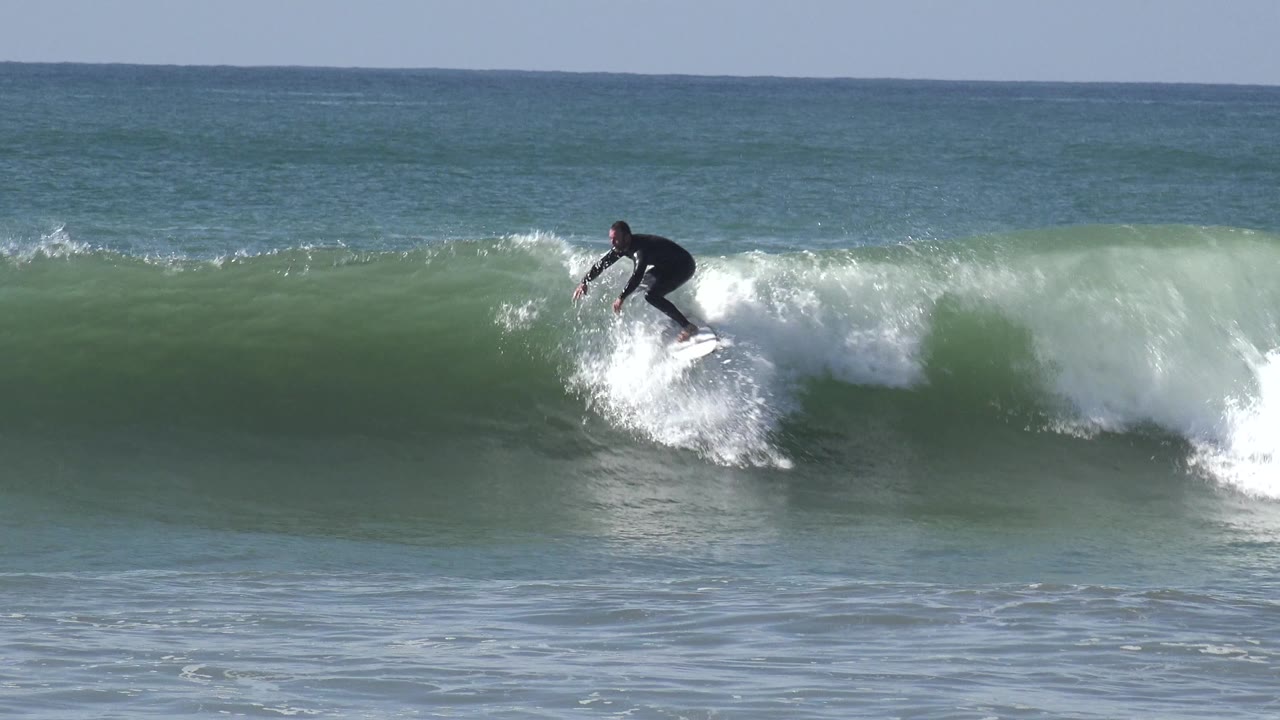 A wetsuited surfer rides down the face of a Huntington Beach wave, with whitewater forming to the right and smooth green water below the board.