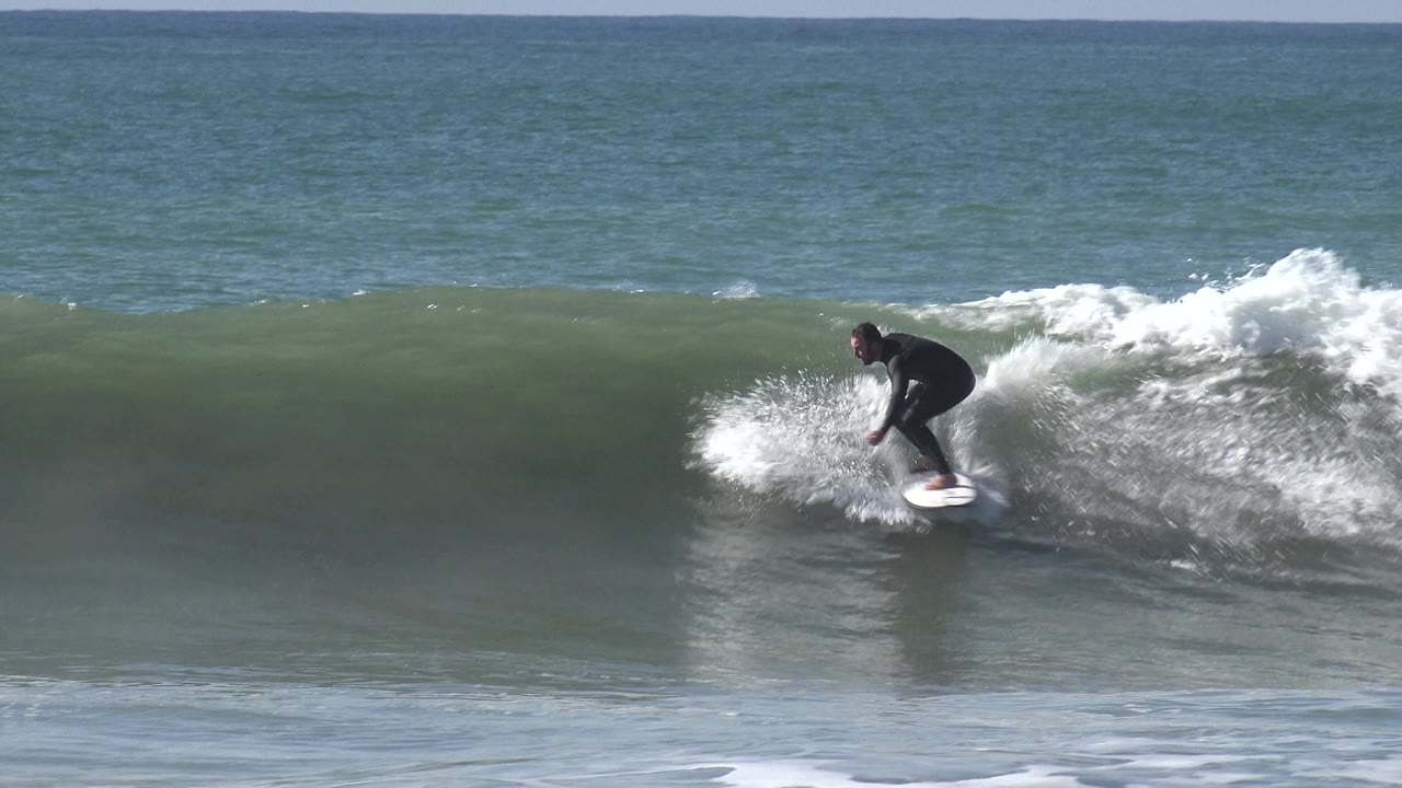At Huntington Beach, California, a surfer in a black wetsuit rides a white surfboard across the face of a green-blue wave as white spray breaks behind him.