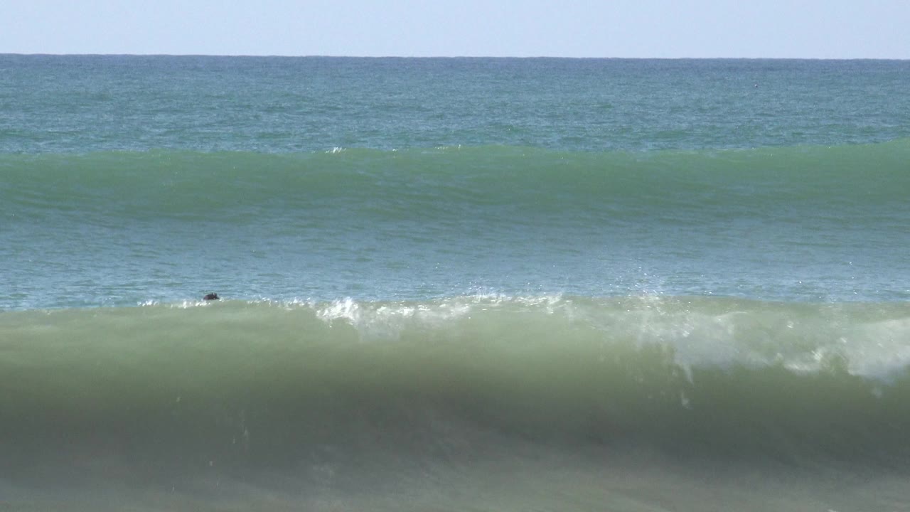 A green-blue swell rises across the frame at Huntington Beach, California, with the ocean horizon high in the background and no visible people in this moment.