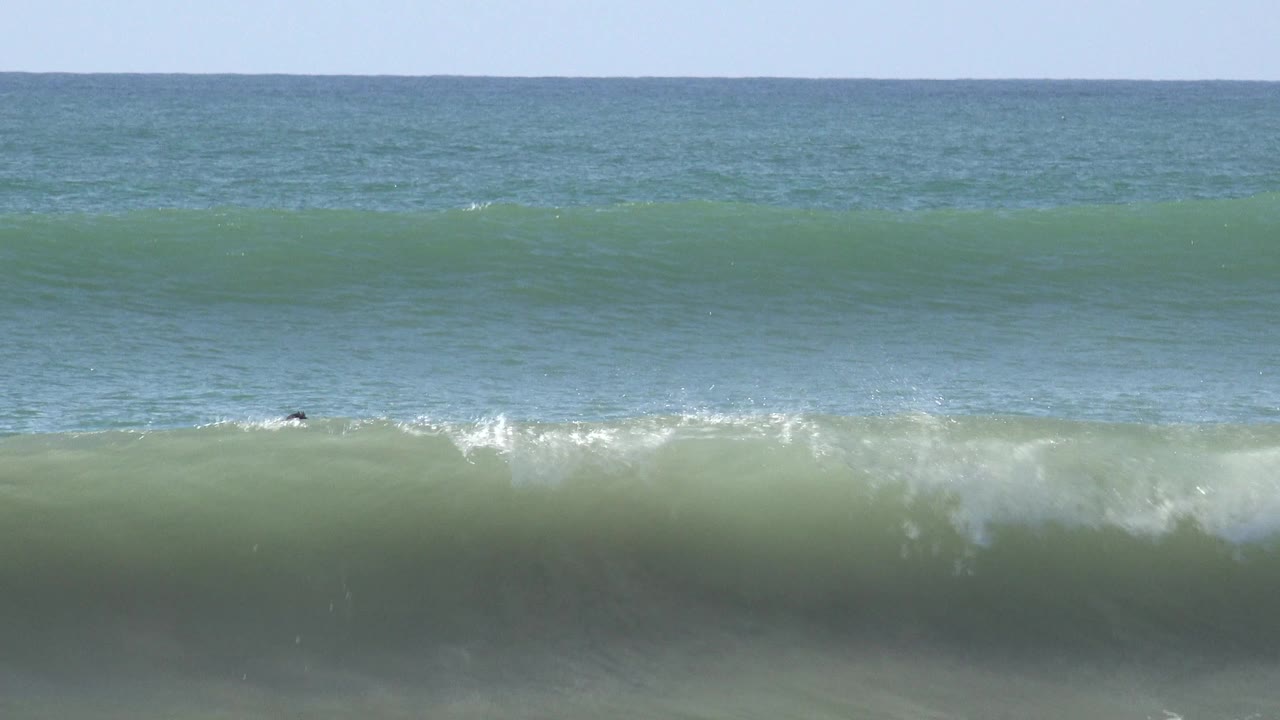 A steep green wave face rolls at Huntington Beach, California, with tiny dark surfer silhouettes near the crest and blue ocean beyond.
