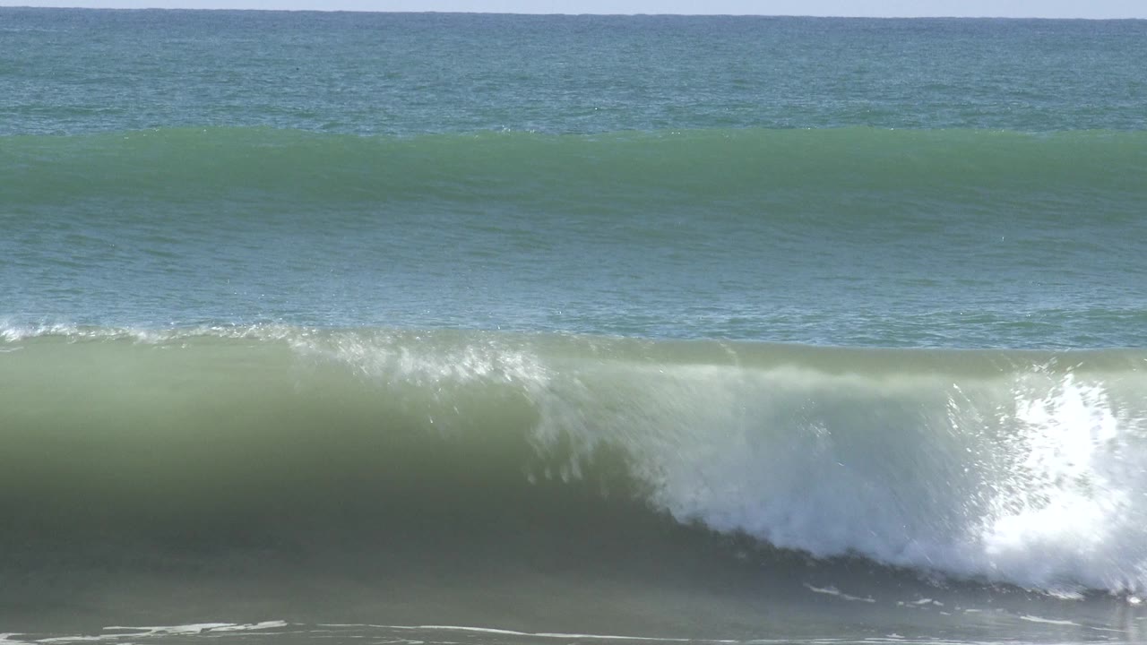 A green wave breaks from the right at Huntington Beach, California, with bright white spray and foam cutting across the wave face and no clearly visible people.