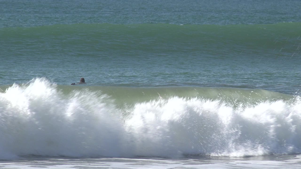 Foamy shore break in the foreground with distant surfers beyond it.