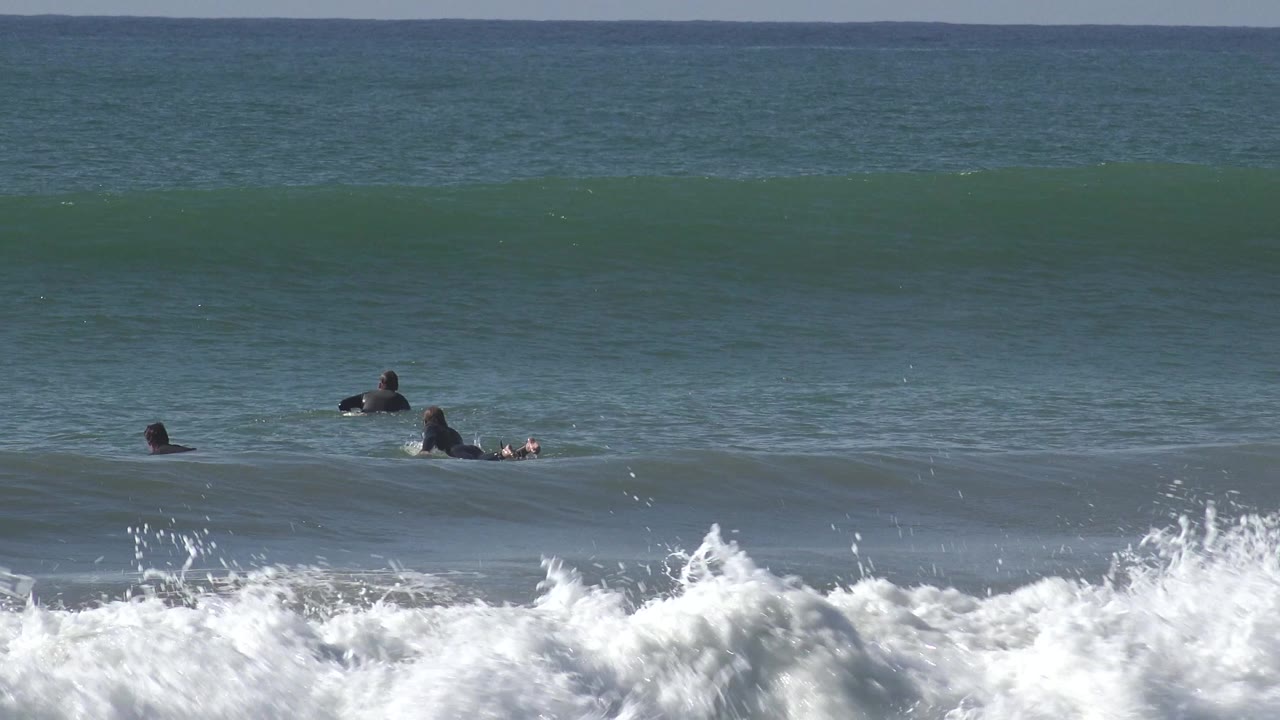 Three surfers on surfboards sit and paddle beyond foamy shorebreak at Huntington Beach, California, with a green wave rising behind them.
