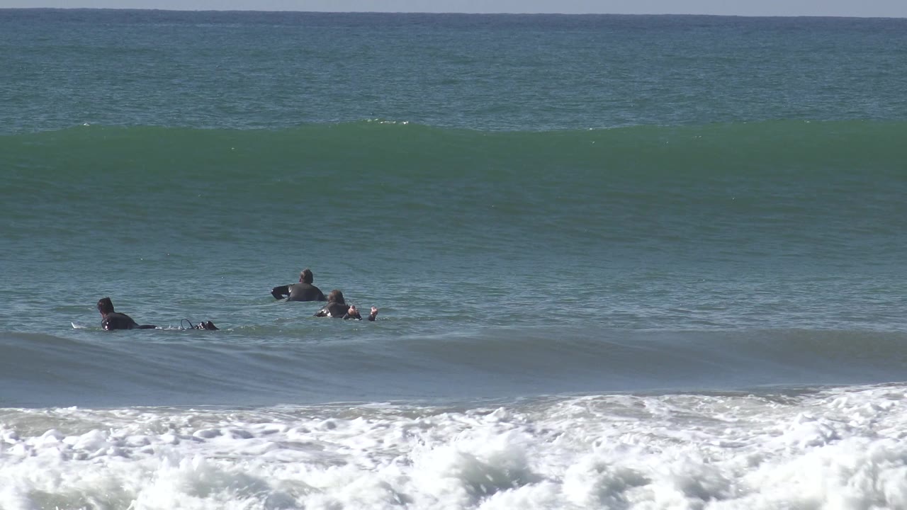 Surfers on boards are visible beyond a band of crashing whitewater at Huntington Beach, California, while a broad green wave line stretches across the frame.