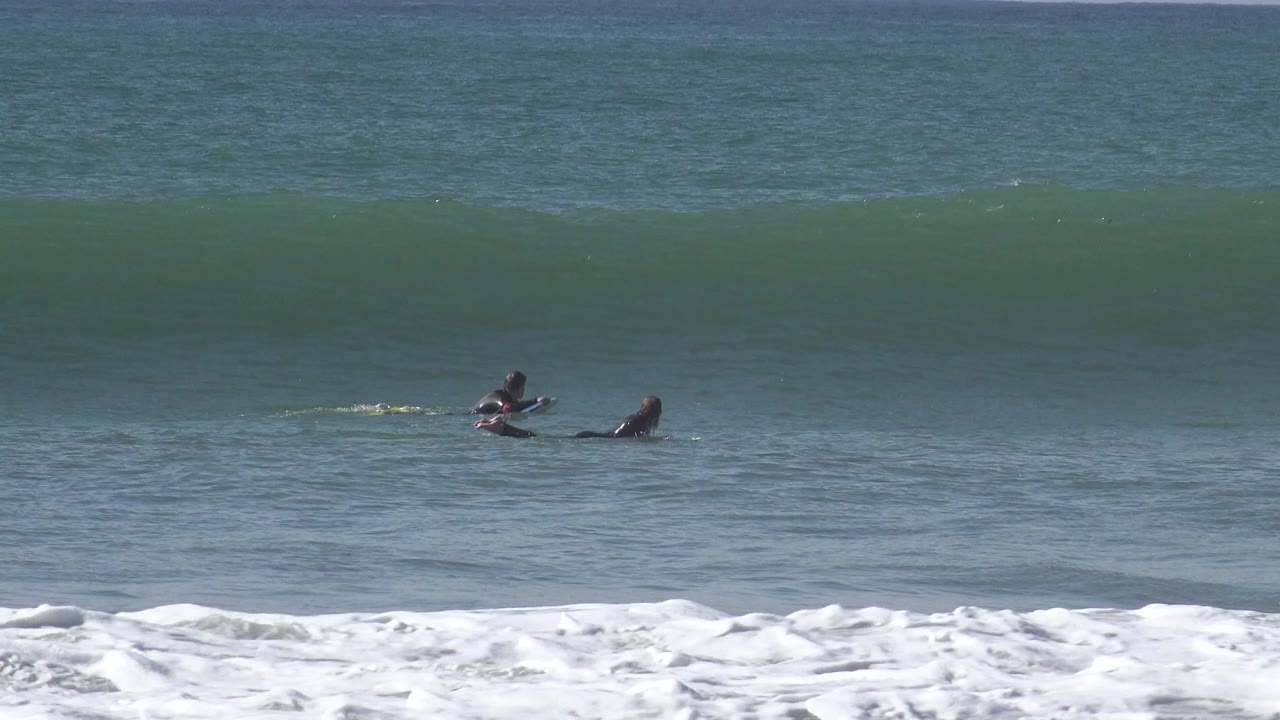 At Huntington Beach, two distant wetsuit-clad people lie on boards as a broad green wave rises behind them and white foam lines the lower frame.