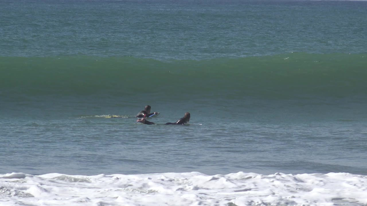 Two board riders remain low in the water as the wave face grows behind them.