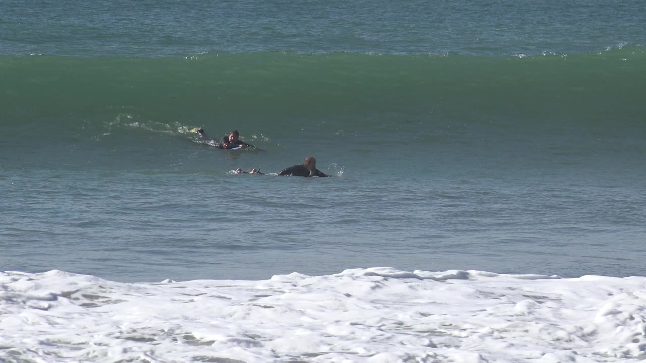 At Huntington Beach, several distant people in dark wetsuits paddle across the blue-green water as the crest of a large wave starts to feather behind them.