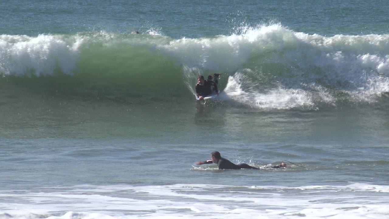 At Huntington Beach, California, a wetsuited surfer sits near the face of a green wave while another person paddles in the foreground surf.