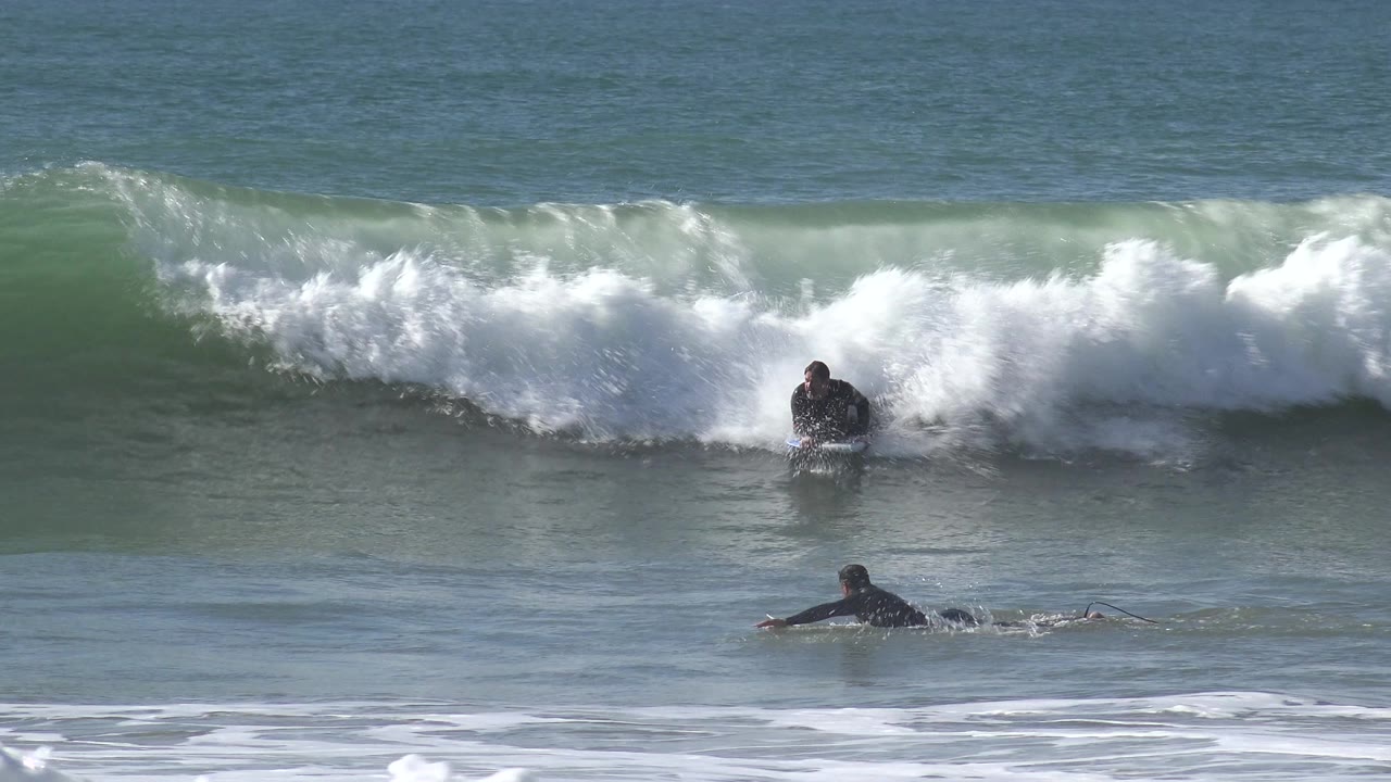 At Huntington Beach, California, a breaking wave throws whitewater across the frame while two wetsuited people remain visible near the impact zone.