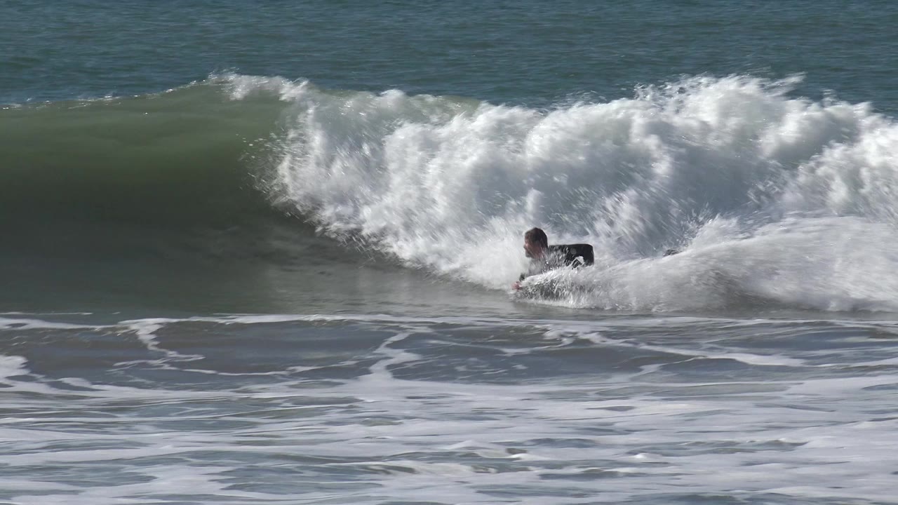 At Huntington Beach, California, a surfer in a dark wetsuit is partly covered by whitewater as a green wave breaks across the midground.