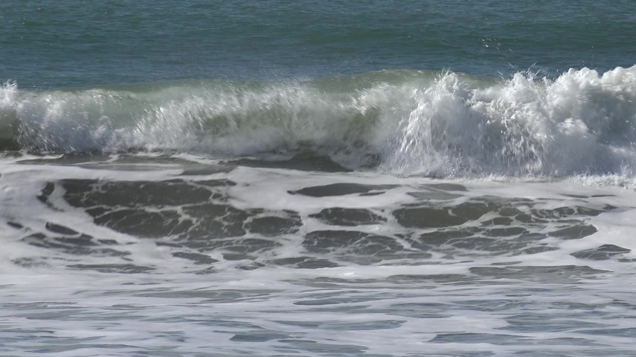 At Huntington Beach, California, a broken wave rolls across the frame with white foam in the foreground and no visible people in this frame.