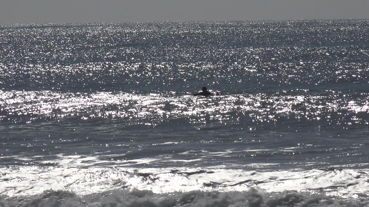 Huntington Beach ocean water sparkles under bright light, with foamy surf near the bottom and tiny distant people or surfers visible offshore.