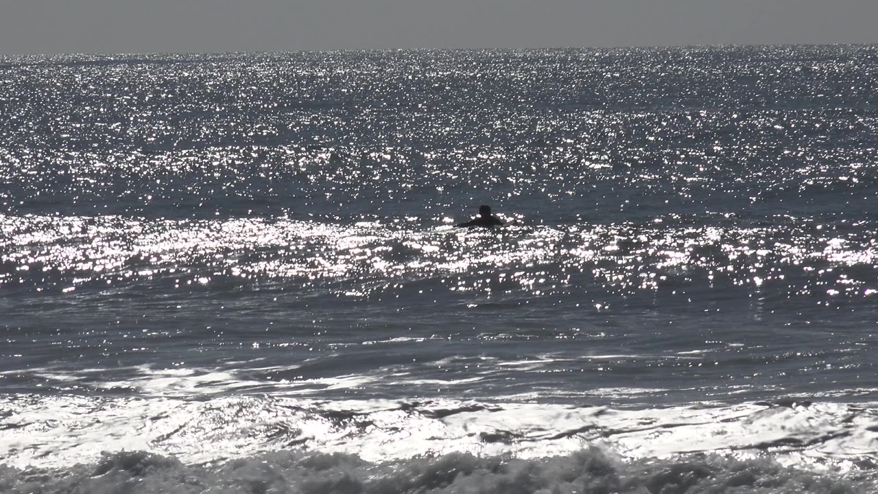Bright Huntington Beach surf fills the frame, with white foam in the foreground and a small dark person or surfer visible beyond the breaking wave.