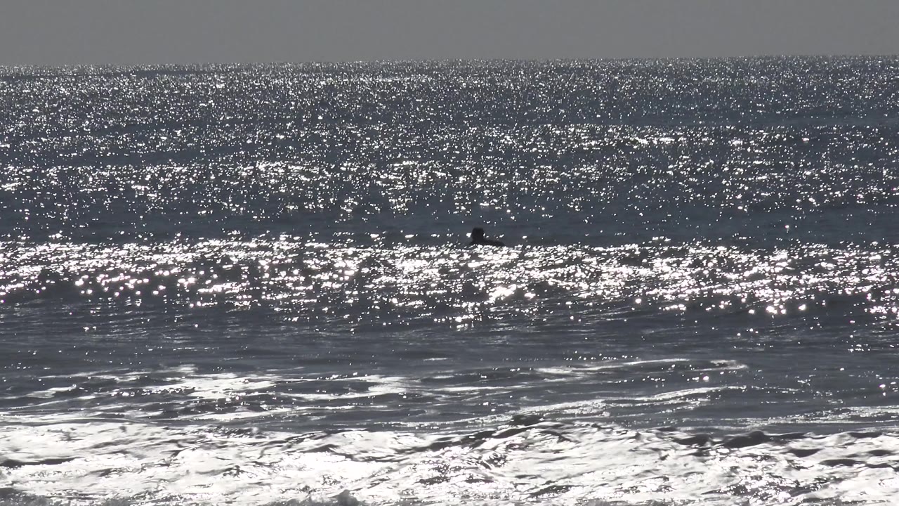 Huntington Beach ocean glare spreads across the upper water while foamy surf rolls below and tiny dark offshore figures remain near the horizon area.