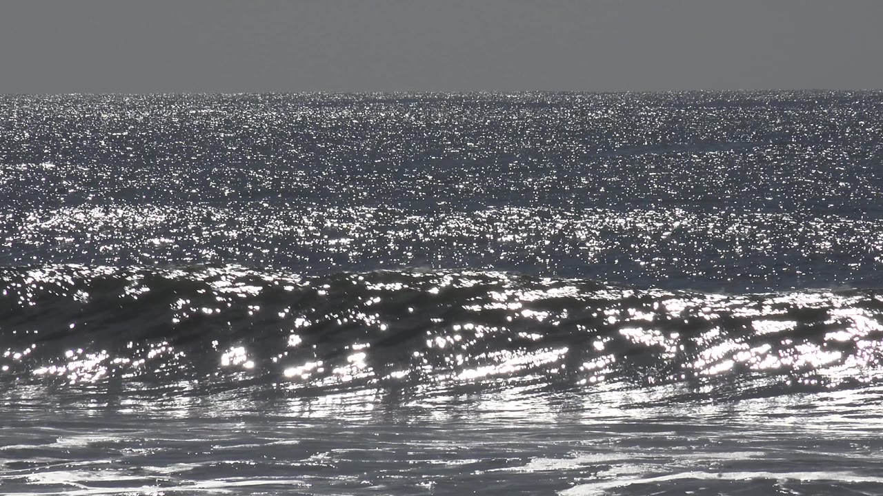 A rolling Huntington Beach wave catches silver sunlight across the water, with foamy surf at the bottom edge and no visible people.