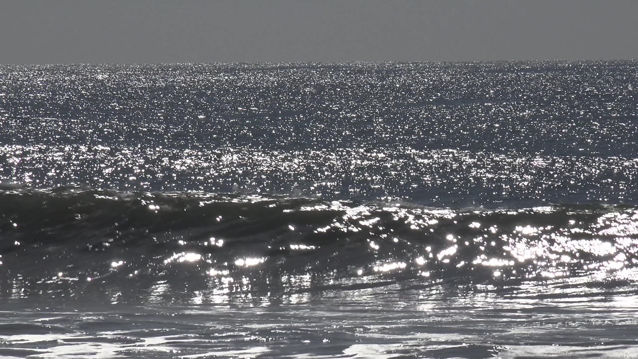 Sun glare scatters across the Huntington Beach ocean while a lower wave breaks near the foreground, with no visible people in this moment.