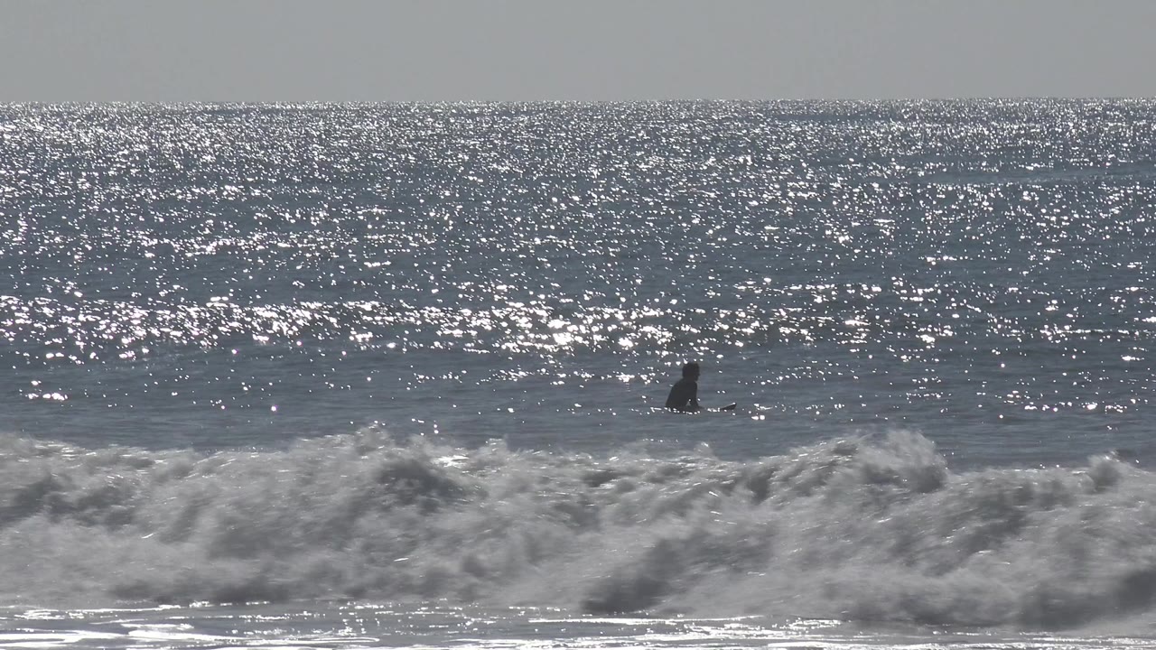 A distant person in dark surf gear sits beyond a breaking wave at Huntington Beach, California, with bright glare scattered across the blue gray ocean.