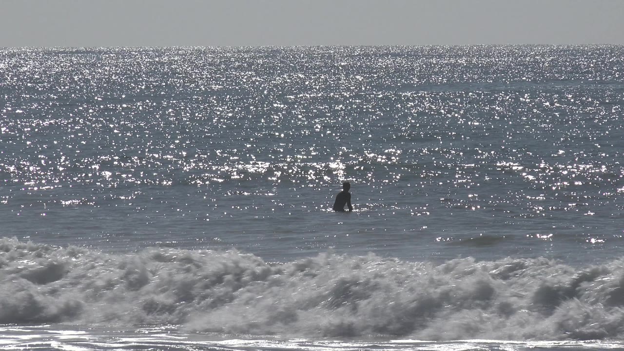 A distant person appears just above the surf line at Huntington Beach, California, with choppy white foam in the foreground and glittering water behind.