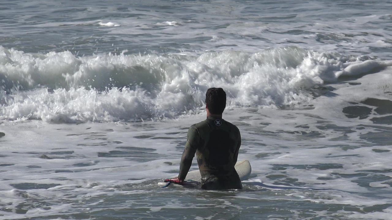 A surfer in a dark wetsuit sits on a surfboard in Huntington Beach, California, facing a line of breaking whitewater.