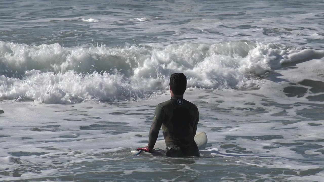 The surfer remains seated on the board in Huntington Beach, California, with more foam spreading across the water in front of the breaking wave.