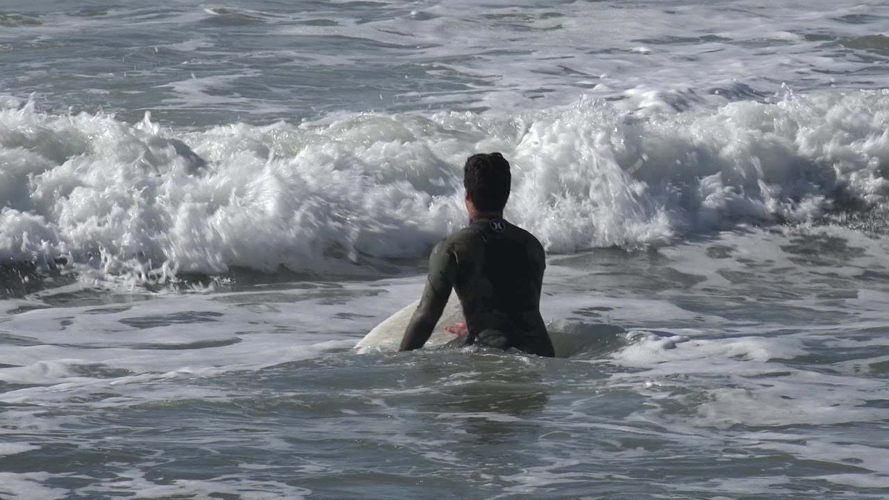 A wetsuited surfer sits partly upright in Huntington Beach, California surf as white foam surrounds the board and a wave collapses behind him.