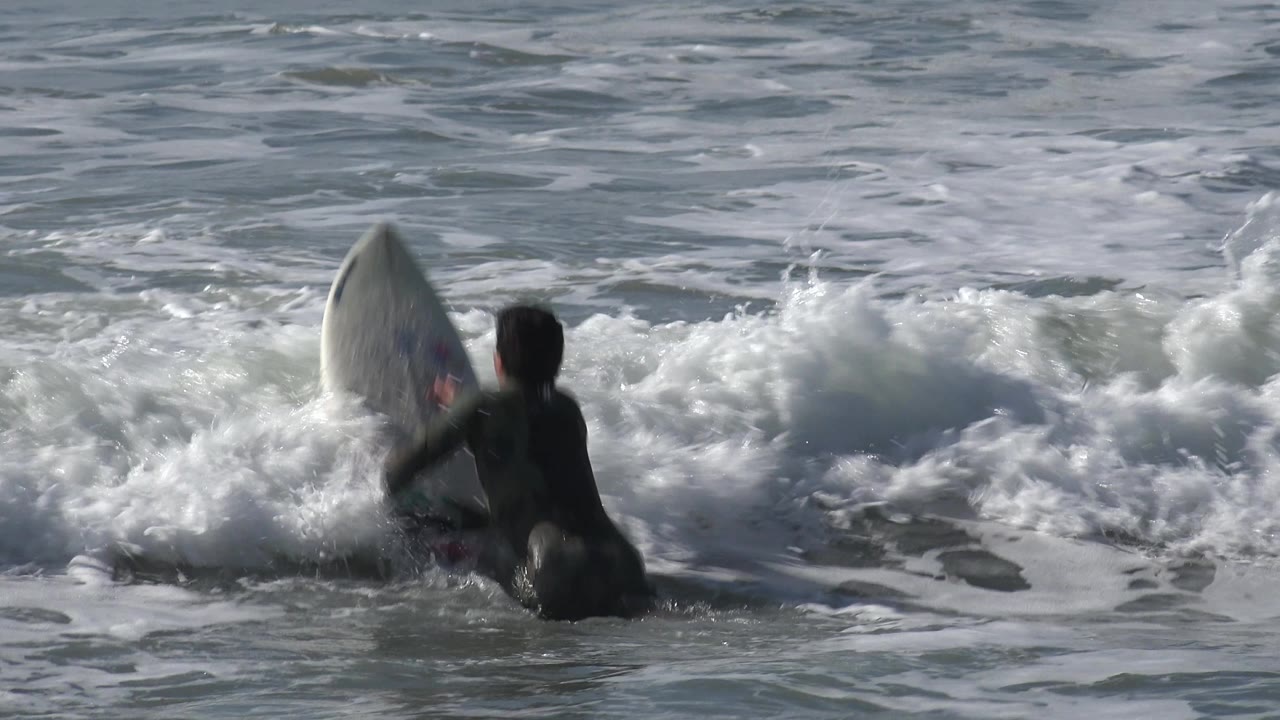 Surfboard pitches upward beside surfer in foamy water.