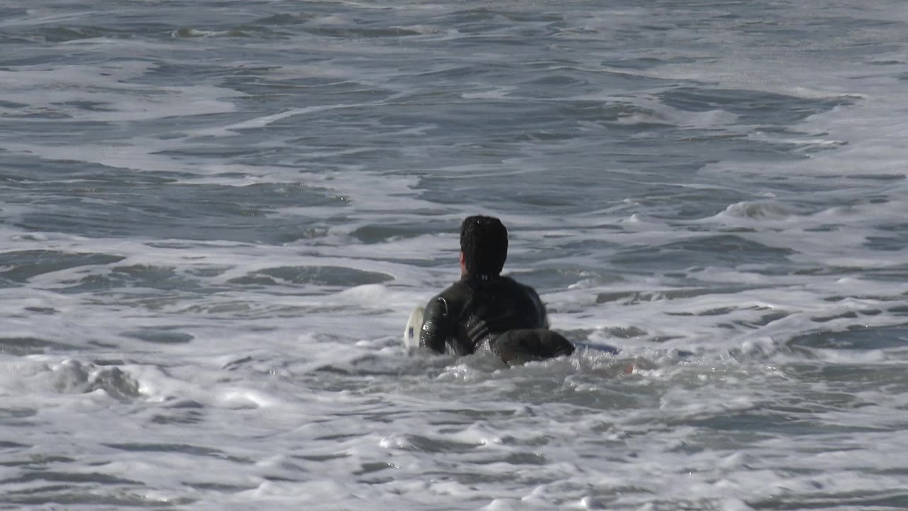 A person in a dark wetsuit paddles away from camera through white surf foam in Huntington Beach ocean water.