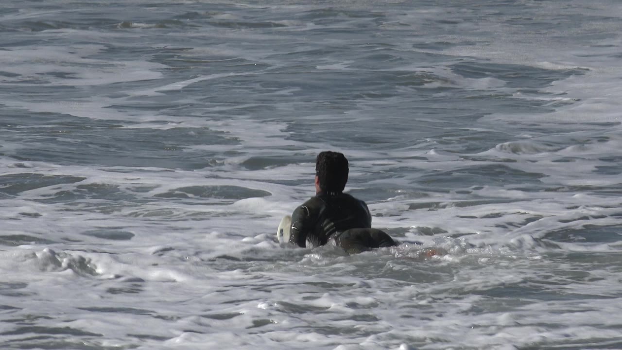 The swimmer remains centered from behind in gray-blue Huntington Beach seawater as a band of foam spreads across the frame.