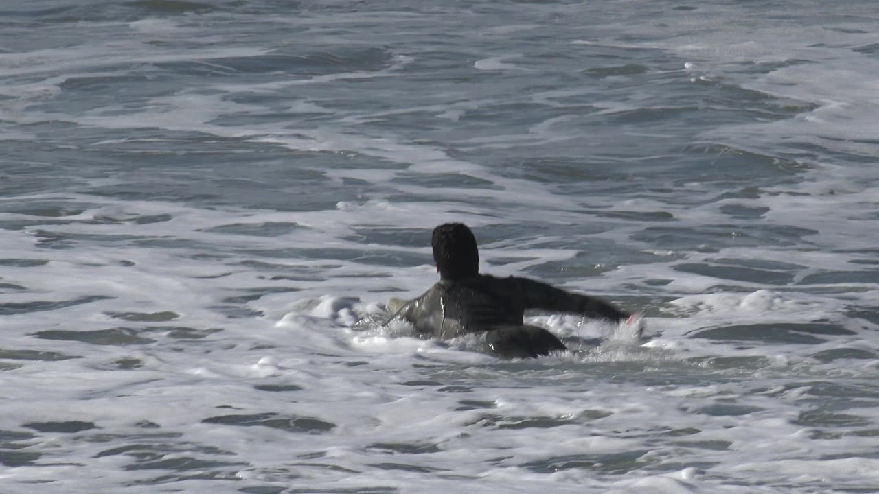 A person in a dark wetsuit paddles slightly to the right through foamy Huntington Beach ocean water, with arms low near the surface.