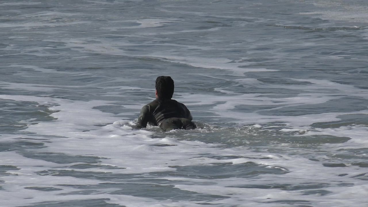 A person viewed from behind paddles through gray-blue surf at Huntington Beach, California, with white foam spread across the water ahead.