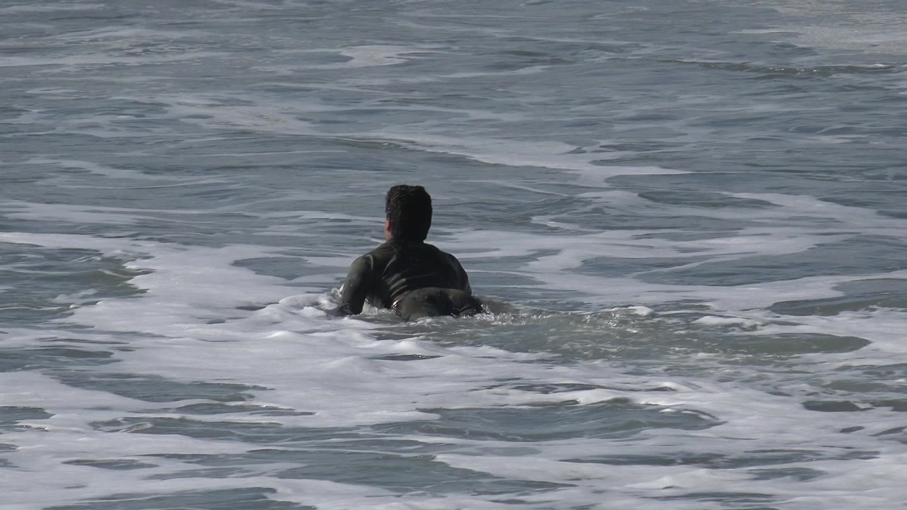 The swimmer remains centered from behind in Huntington Beach surf as bands of white foam drift across the gray water.