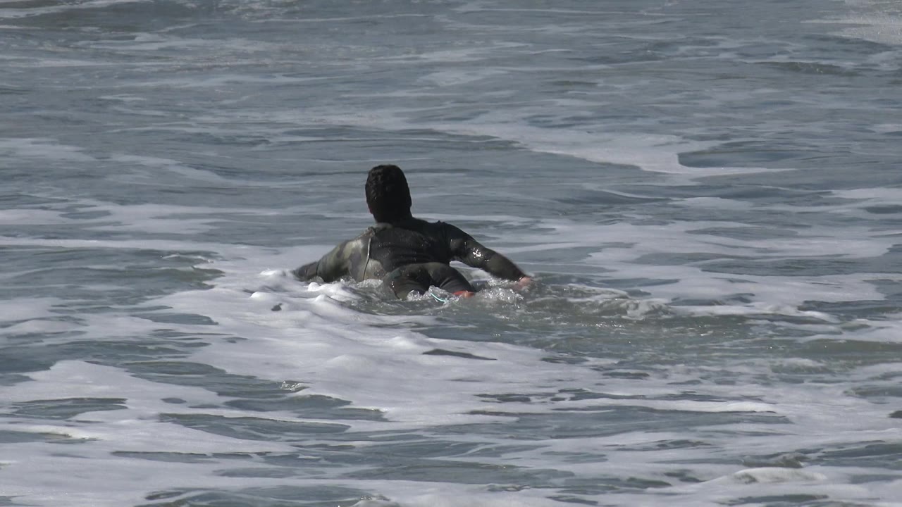 A person in dark clothing or swimwear paddles away from camera in Huntington Beach water, with foamy surf surrounding the body.