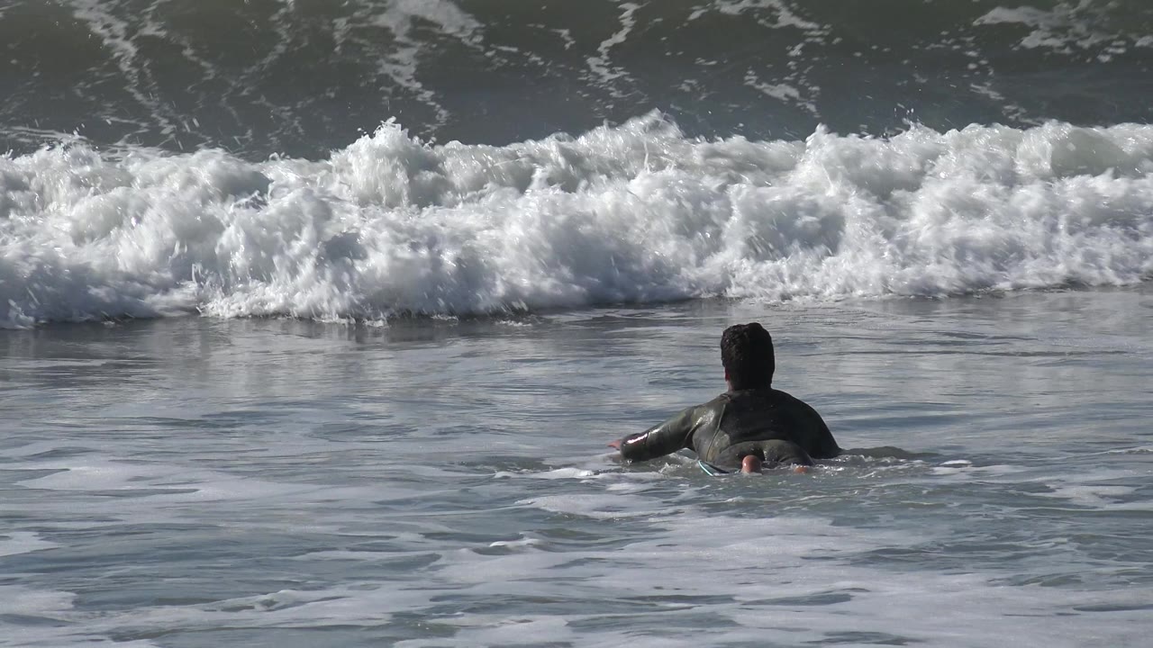 A surfer in dark gear paddles away from camera on a surfboard toward a white breaking wave at Huntington Beach, California.