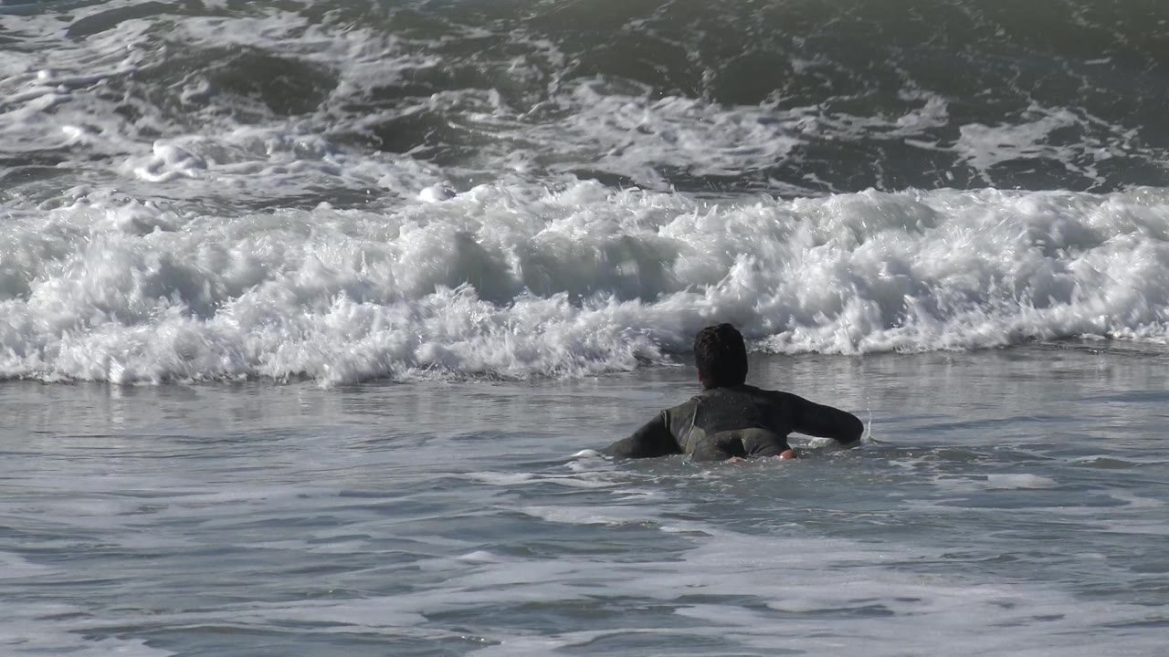 A surfer paddles slightly right of center through rippled ocean water as the whitewater builds behind him at Huntington Beach, California.