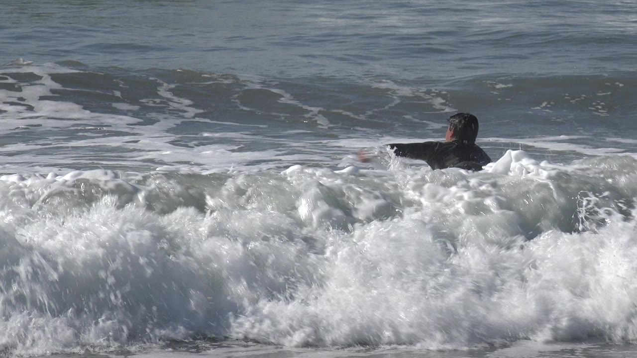 A surfer in dark swimwear paddles through foamy Huntington Beach surf as a breaking wave fills the lower frame.